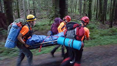 Three people in helmets and rescue gear carry a rescue litter through the forest
