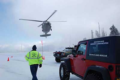 A helicopter flies close to the ground with snow on the ground, a Jeep in the foreground, and a person walking in a bright green jacket