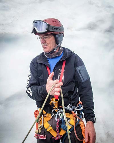 A person with glasses and a mustache in a harness and wearing a helmet with ski goggles attached holds a rope between index and middle fingers on a snowy slope